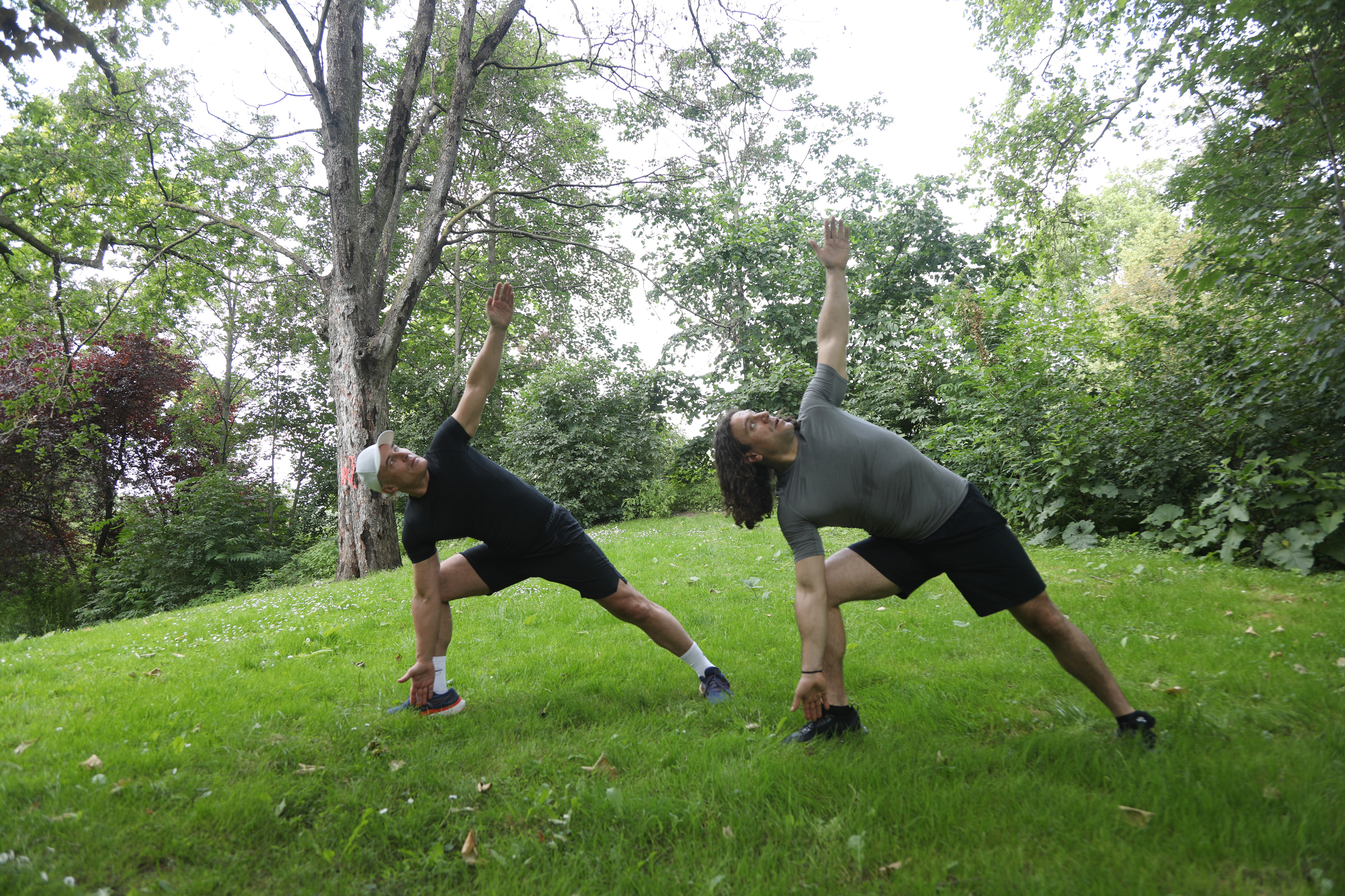Ejercicios de Yoga al Aire Libre - Estiramiento Dos hombres realizan ejercicios de estiramiento al aire libre, en un campo verde, con árboles al fondo.