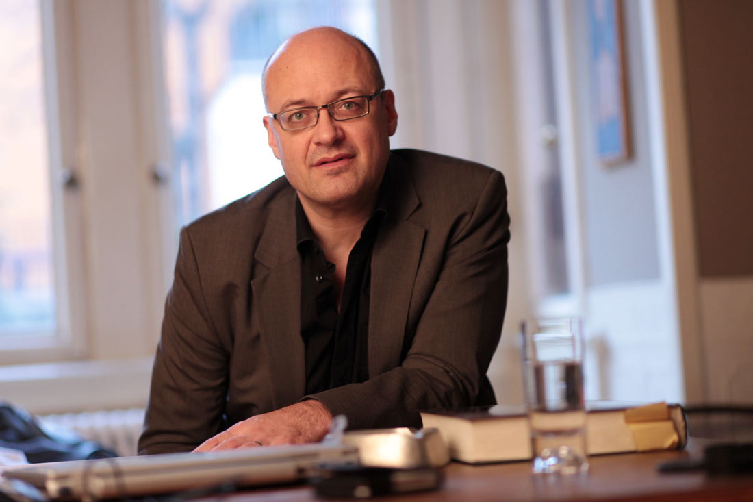 Un hombre en traje sentado a la mesa con un libro y un vaso de agua en una habitación luminosa.
