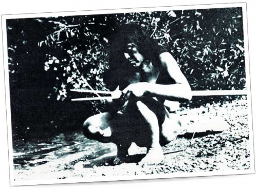 Un hombre sentado en la orilla de un río trabaja en una lanza. Está descalzo y lleva ropa natural.