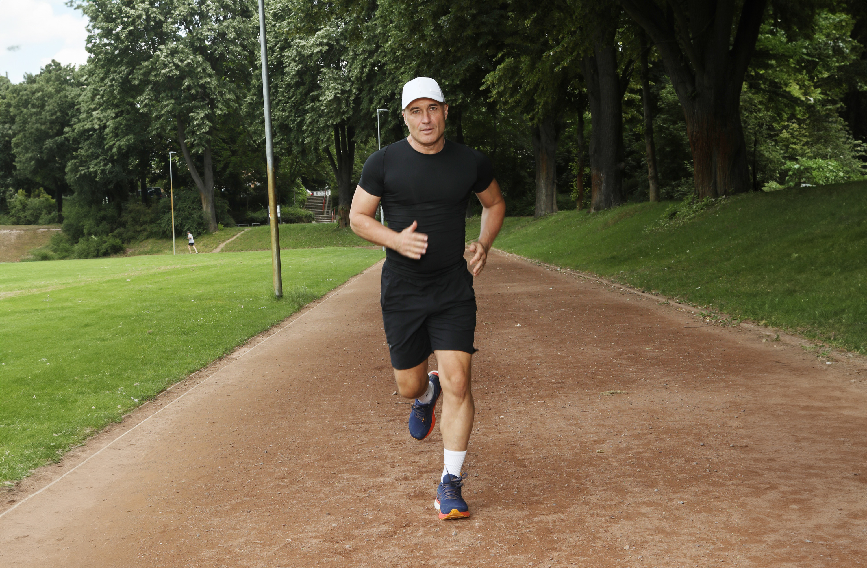 Zapatillas de correr para hombres – Modelos deportivos de 10 km Hombre corre por un sendero forestal, vestido con ropa deportiva negra y zapatillas de correr azules.