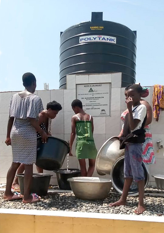 Tanque de agua – Extracción de agua en recipientes Grupo de niños extrayendo agua de un tanque, varios recipientes, ropa clara, entorno soleado.