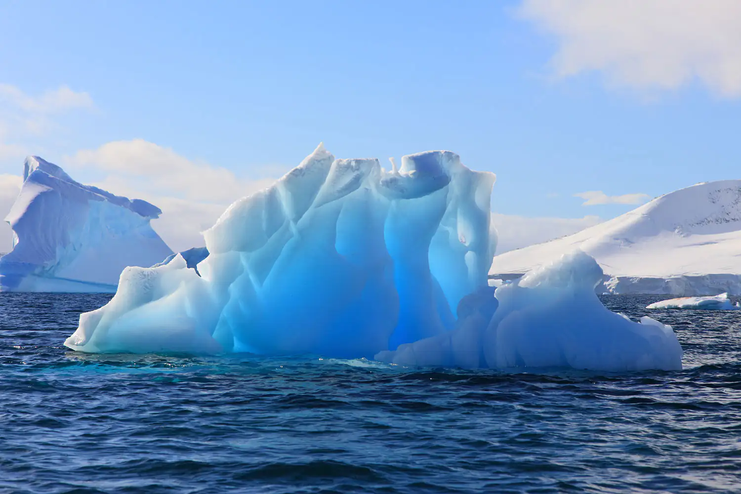 Un iceberg azul flota en aguas tranquilas, rodeado de montañas nevadas y cielo.