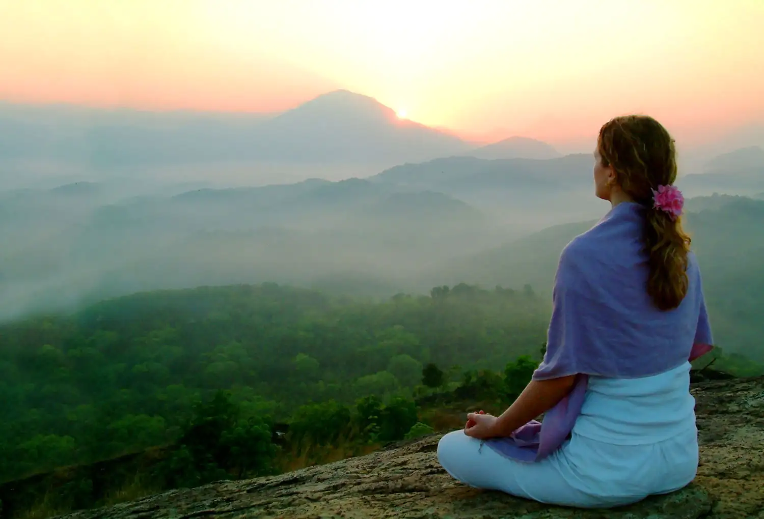 Mujer sentada meditando sobre una roca, rodeada de un paisaje montañoso nublado al amanecer.