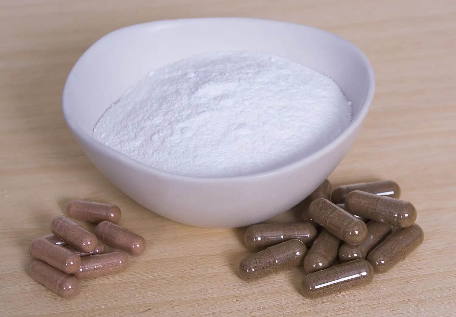 Polvo de creatina blanca en un tazón, junto a cápsulas marrones, sobre una superficie de madera.