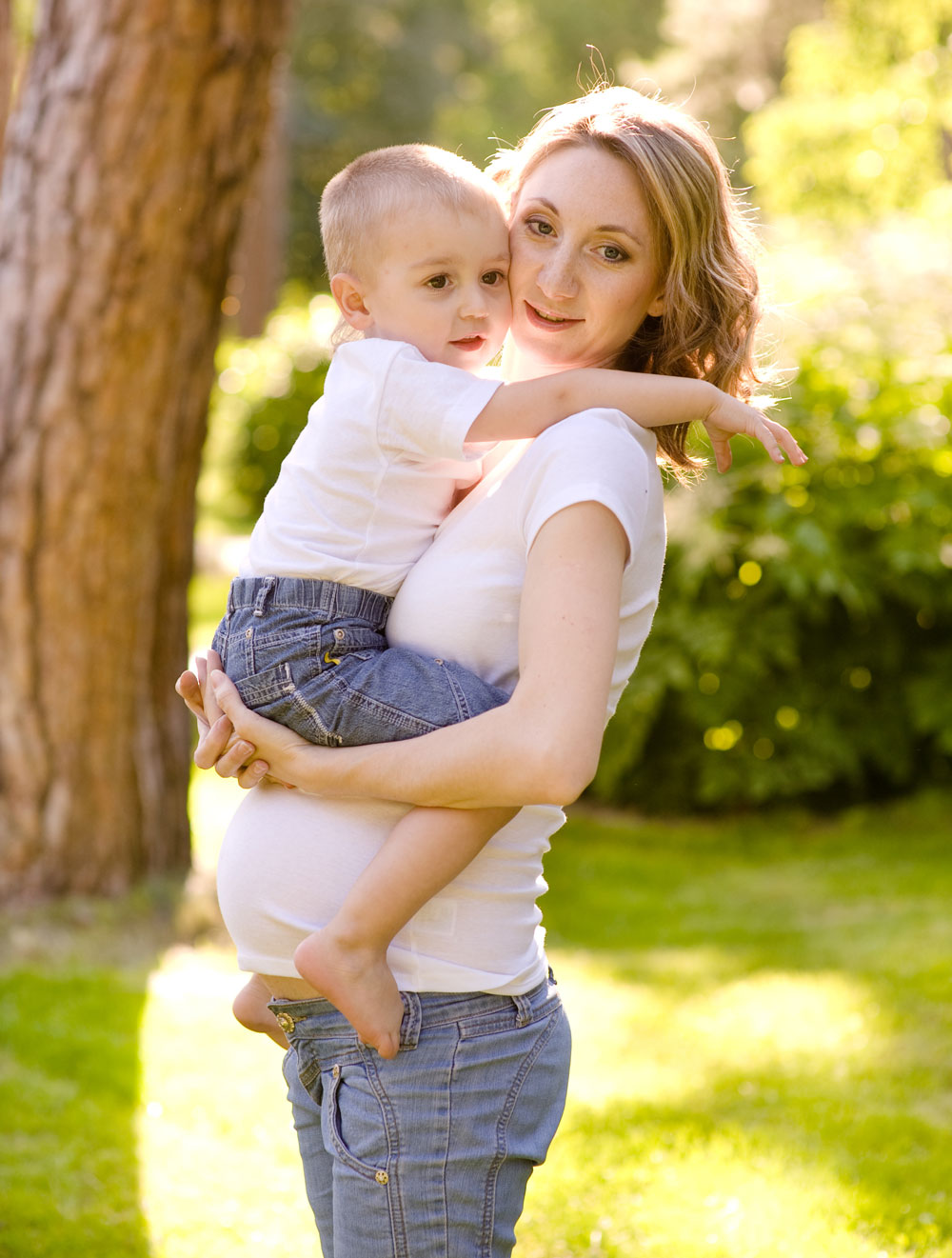 Una mujer embarazada sostiene a un niño en brazos, al aire libre, en un entorno verde.