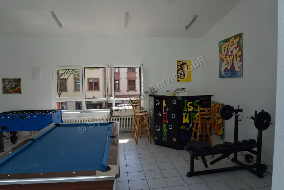 Sala de entretenimiento con mesa de billar y equipamiento de fitness Sala de entretenimiento luminosa con mesa de billar, futbolín, equipo de fitness y barra, gran ventana frontal.